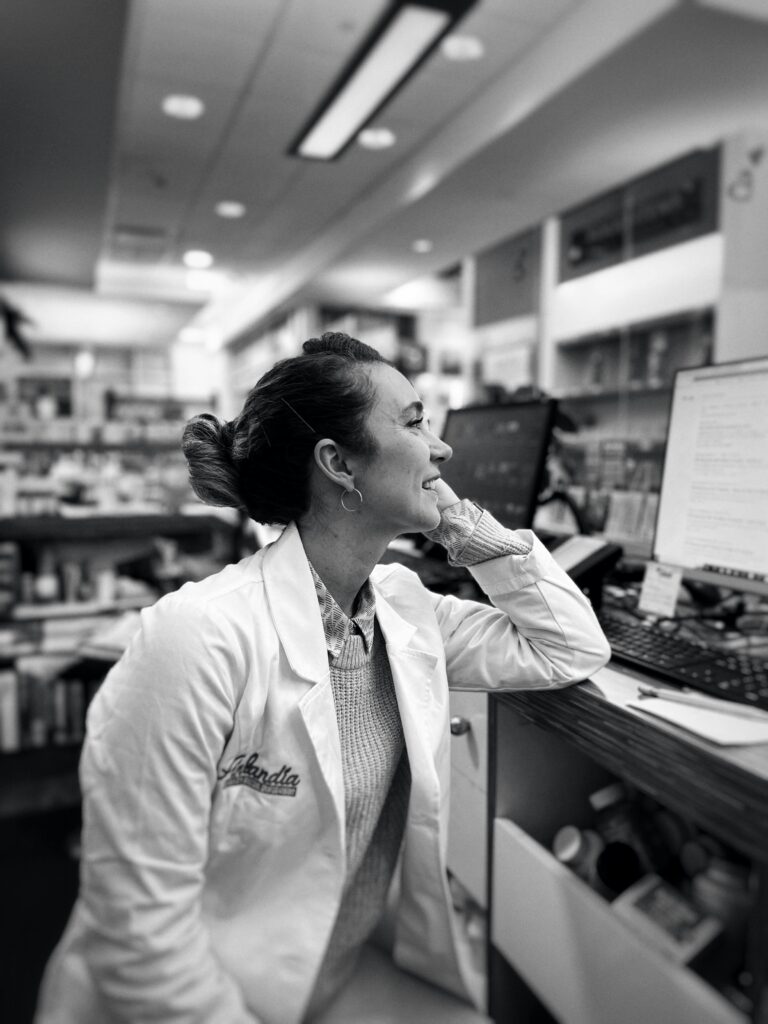 A smiling female pharmacist in a white coat working at a computer in a pharmacy.