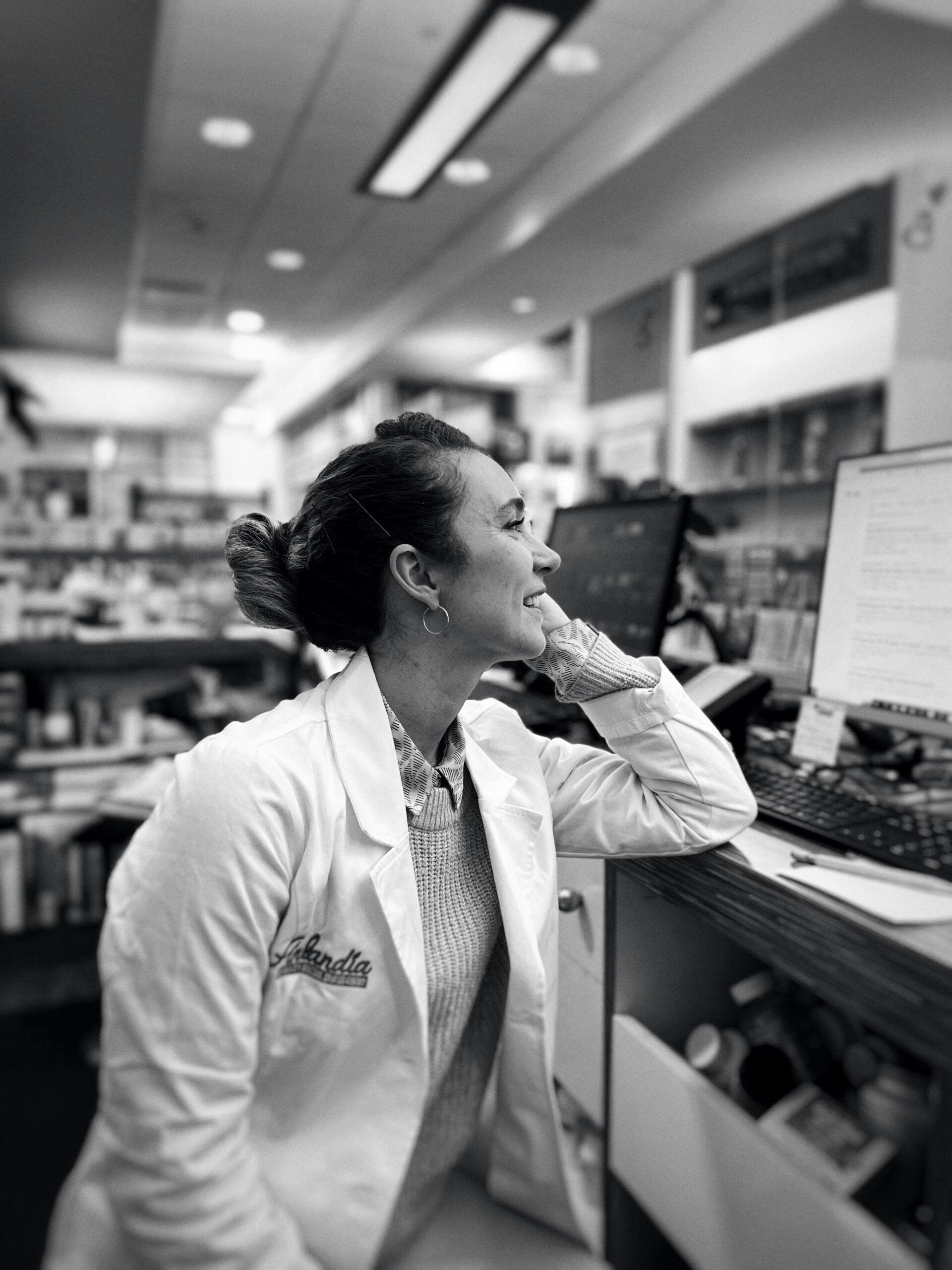 A smiling female pharmacist in a white coat working at a computer in a pharmacy.